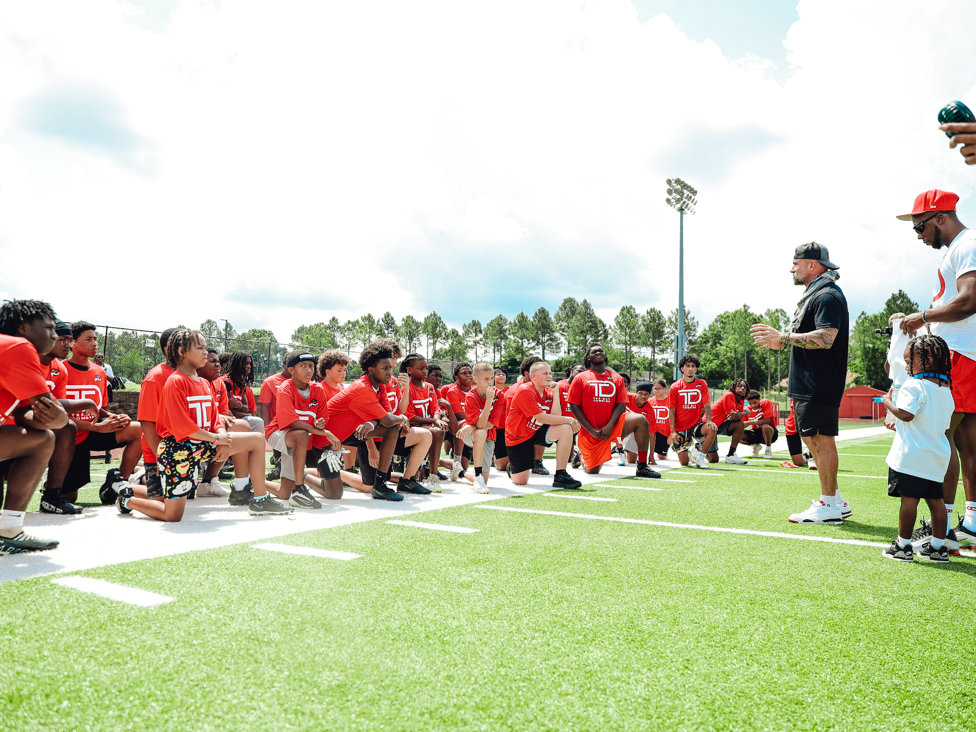 Coach Andy McCloy talking to a huddle of kneeling young athletes in Tre’ Day jerseys on the field