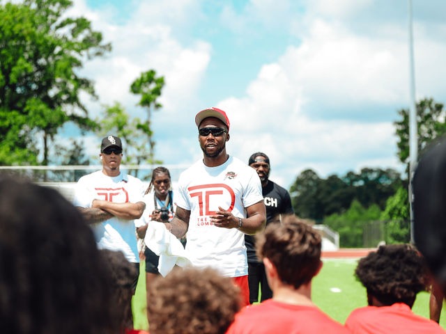 Chester 'Tre'' Rogers speaking to a huddle of young athletes in Tre’ Day jerseys at Milton Frank Stadium